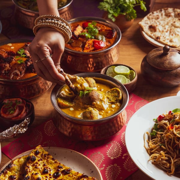 A hand with bangles dips naan into a copper bowl of Indian curry, surrounded by other dishes.